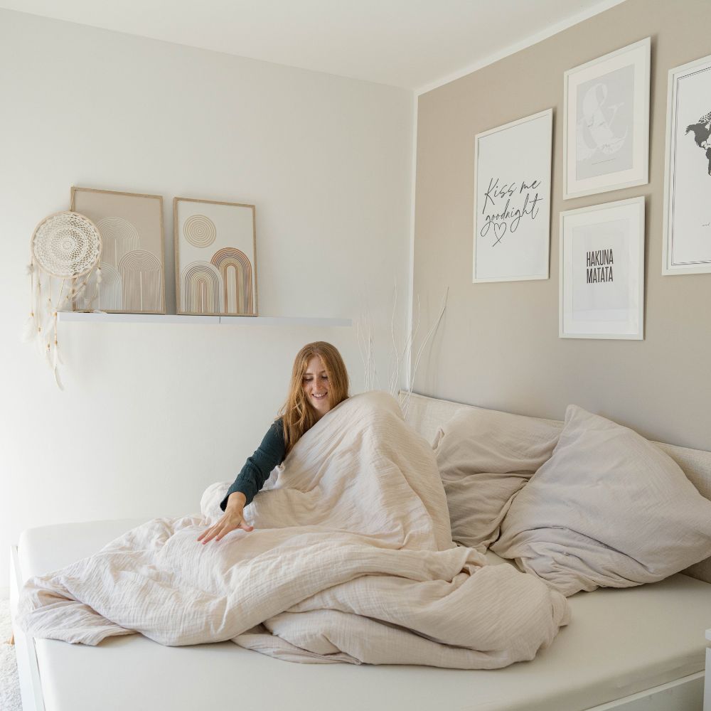 Cozy Set: Frau sitzt im Bett mit cremefarbener Musselin-Bettwäsche im hellen Schlafzimmer