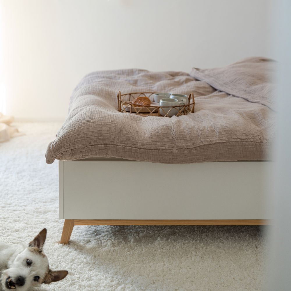Cozy Set: Bett mit taupefarbener Musselin-Bettwäsche und Frühstückstablett, davor ein kleiner Hund