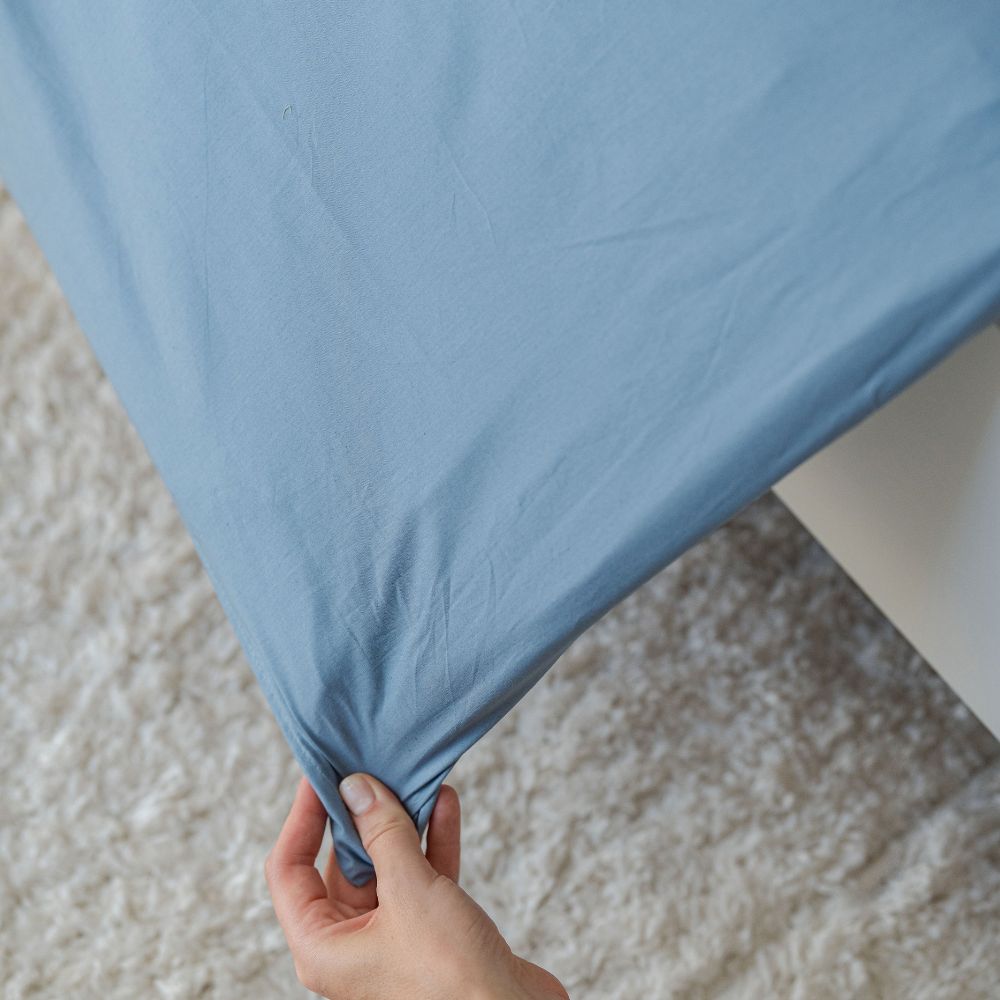 Hand hält die Ecke eines nebelblauen Perkal-Deckenbezugs über hellem Teppich