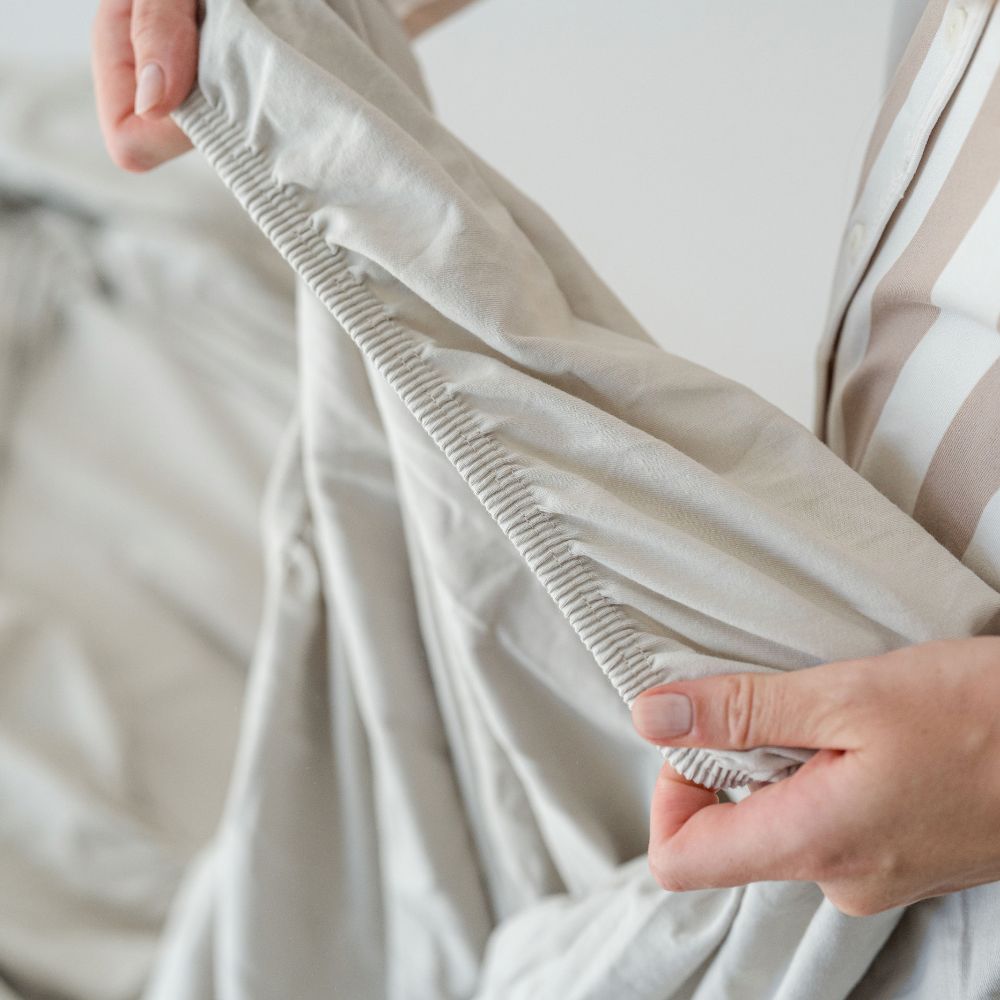 Hand zeigt den elastischen Rundum-Gummizug eines salbeigrauen Spannbettlakens aus Bio-Baumwolle