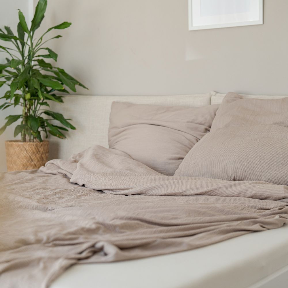 Bett mit taupefarbener Struktur-Bettwäsche aus Bio-Baumwolle, daneben eine Zimmerpflanze