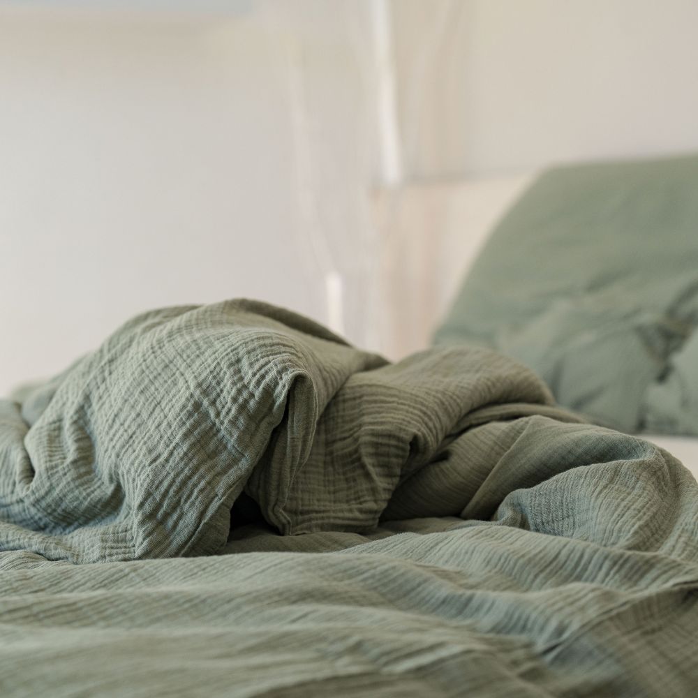 Zerwühlte olivgrüne Musselin-Bettwäsche aus Bio-Baumwolle auf einem hellen Bett