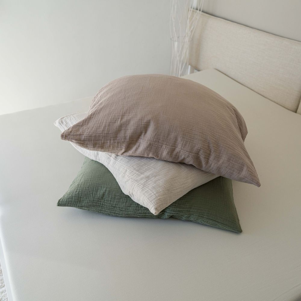 Drei Musselin-Kopfkissen in Taupe, Creme und Olivgrün auf einem weißen Spannbettlaken