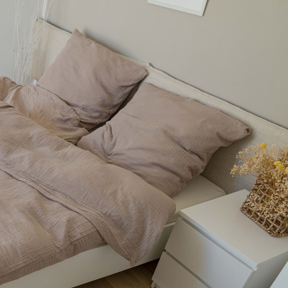 Taupefarbene Musselin-Kopfkissen und Bettdecke auf einem Bett neben weißem Nachttisch